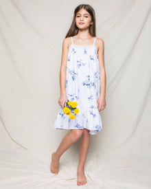 A young girl in Petite Plumes Indigo Floral Twill Lily Nightgown stands barefoot against a light background, holding a bunch of yellow flowers.