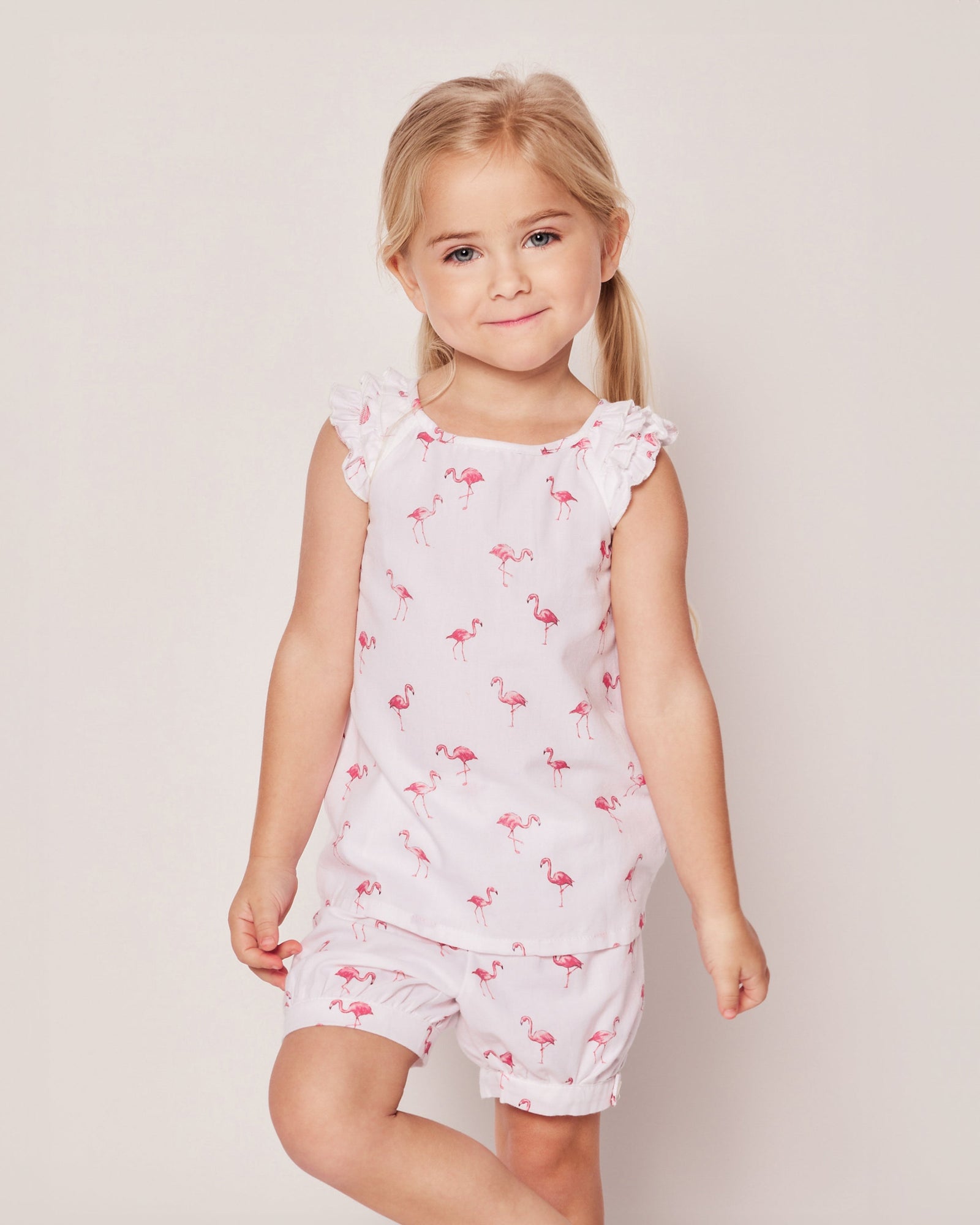 A young blonde girl wears the Girls Twill Amelie Short Set in Flamingos by Petite Plume, featuring a sleeveless top with ruffled shoulders and matching shorts in a playful pink flamingo pattern. She smiles gently against a plain, light-colored background.