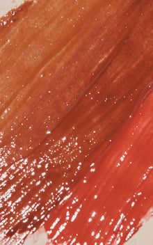 Glossy coral-red lipstick swatch with warm brown undertones and scattered sparkle highlights