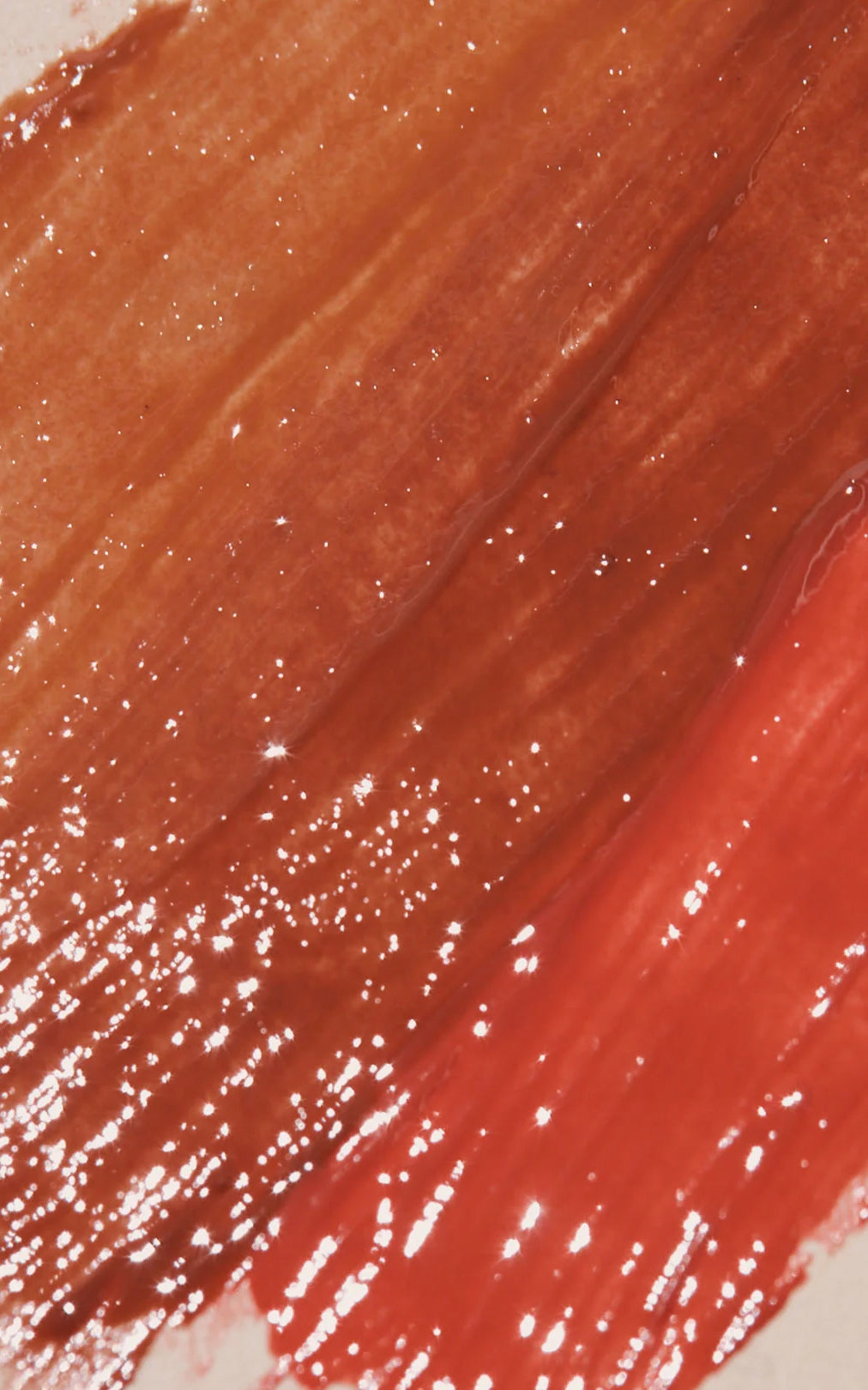 Glossy coral-red lipstick swatch with warm brown undertones and scattered sparkle highlights