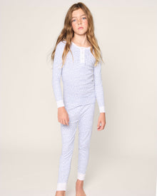 A young girl with long, brown hair poses against a light background in Petite Plumes Kids Pima Snug Fit Pajama Set in Bluehearts. The set features a soft, floral-patterned top and pants. Barefoot, she gazes at the camera with an innocent charm.