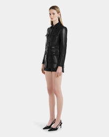 A woman with long blonde hair and a neutral expression wears the Rudsak DANICA - 8125025 Black womens leather jacket, styled with front pockets, black shorts, and black high heels against a plain white background.
