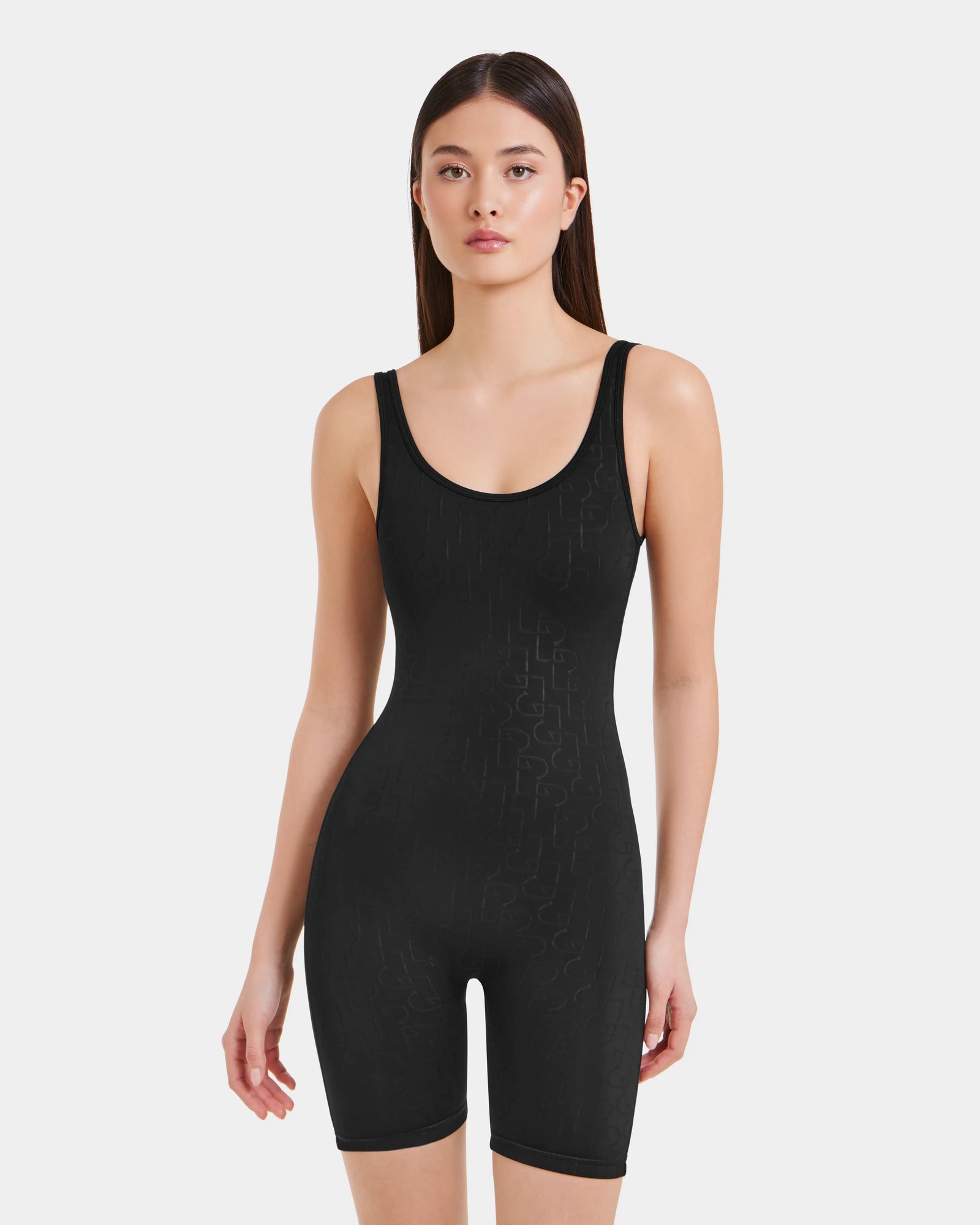 A woman with long, straight brown hair models the Rudsak KAILEY - 8523012 Black Pattern fitted sleeveless bodysuit romper with a scoop neck and mid-thigh length, standing against a plain light background.