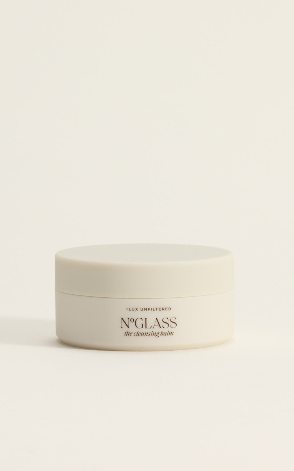 Small white jar labeled 'The Cleansing Balm — LUX UNFILTERED' on a neutral pale background