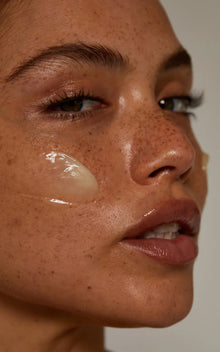 Close-up of The Cleansing Balm on a freckled model's cheek showcasing translucent balm texture on neutral background