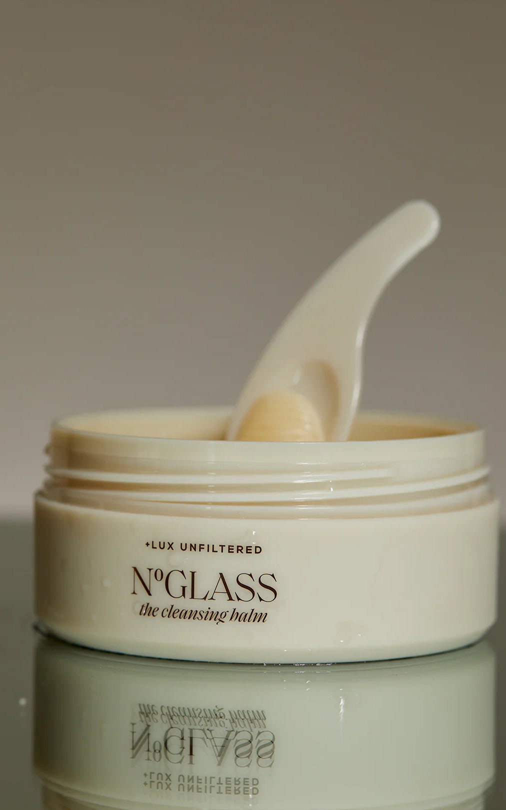 Close-up of open jar of The Cleansing Balm showing cream balm and spatula on a reflective surface