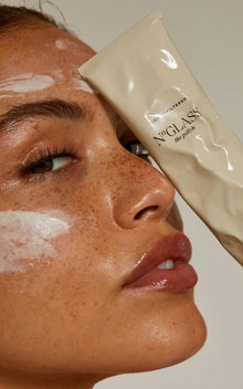 Close-up of freckled woman's face with cream streaks, holding beige tube labeled N°GLASS the polish and LUX UNFILTERED