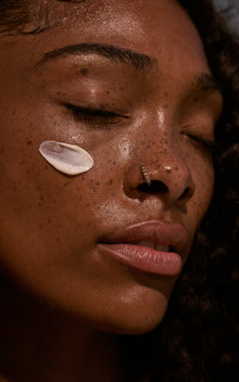 Close-up of a freckled face with a cream dab on the cheek, closed eyes, rhinestone nose hoop, and glossy slightly parted lips.
