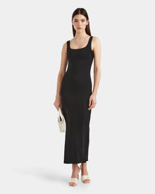 A woman wears the Rudsak WANDA - 8524035 Black fitted sleeveless maxi dress with stretch performance fabric, paired with white high-heeled sandals and a small white woven handbag, against a plain white backdrop.