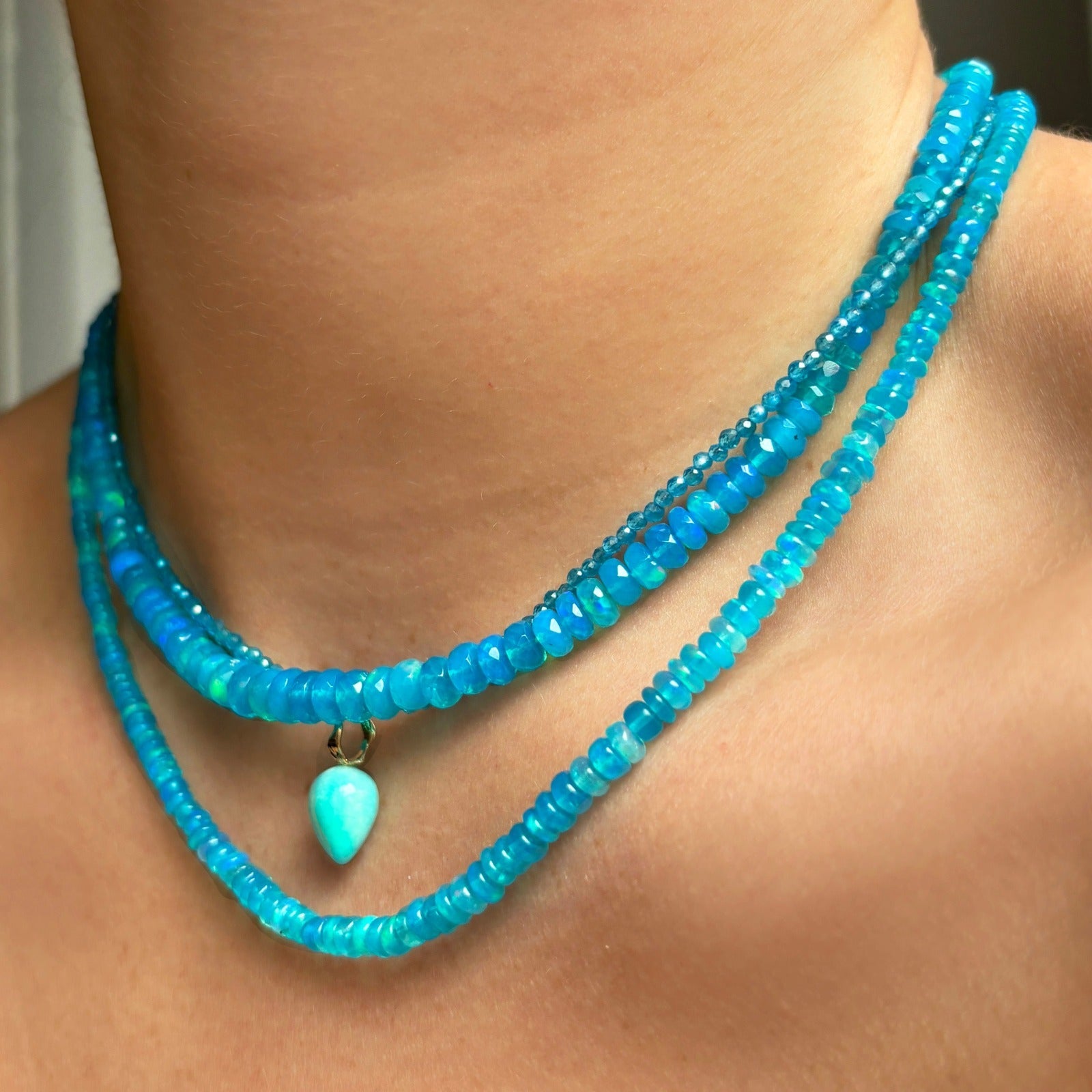 Shimmering beaded necklace made of smooth opal rondels in shades of bright light blue on a slim gold lobster clasp. Styled on a neck layered with a turquoise acorn drop charm.