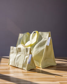 Two light yellow graf lantz canvas bags with short handles sit on a sunlit wooden surface against a gray backdrop, ideal for beach outings. Resembling classic cotton totes, each bag has a small white tag with text.