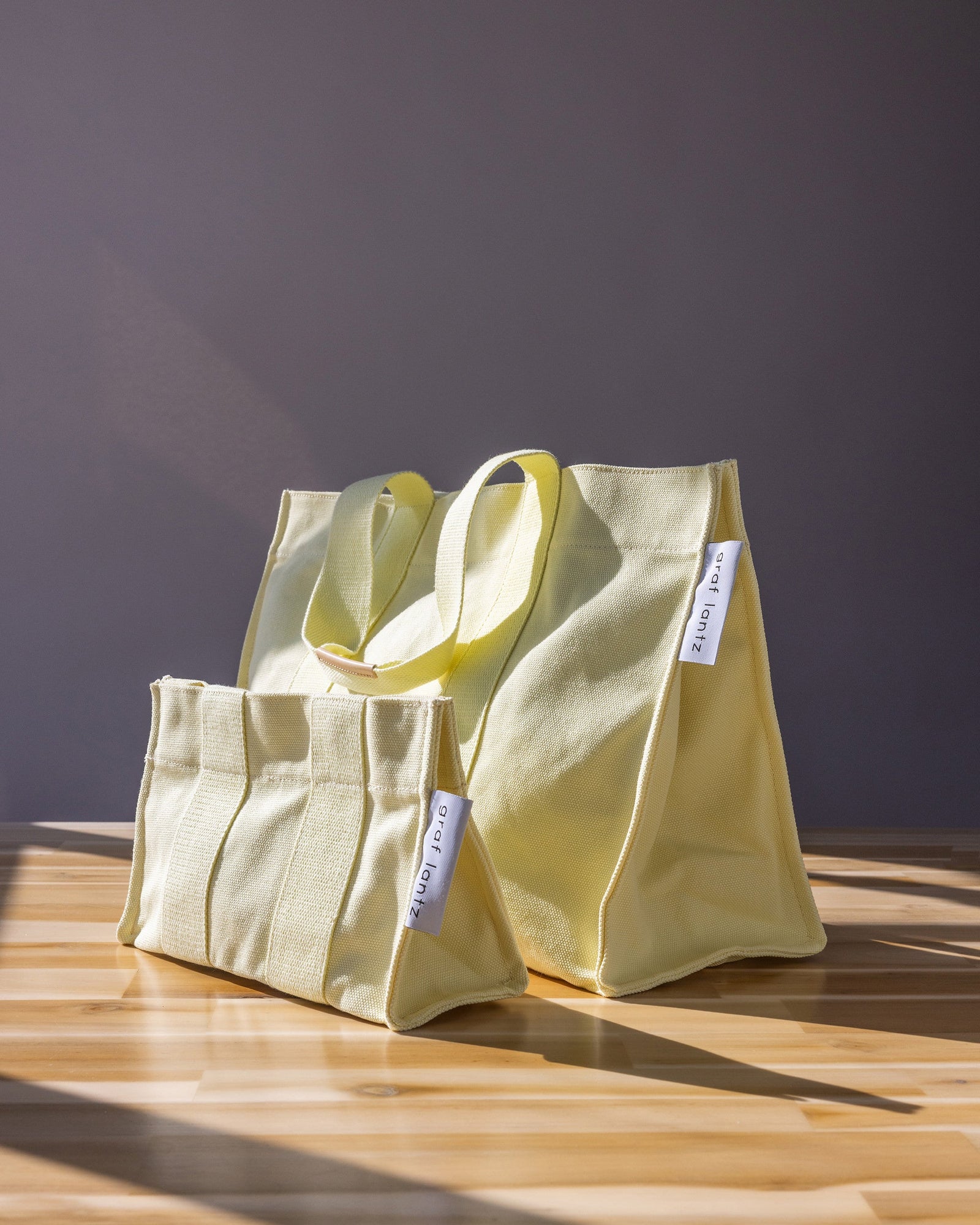 Two light yellow graf lantz canvas bags with short handles sit on a sunlit wooden surface against a gray backdrop, ideal for beach outings. Resembling classic cotton totes, each bag has a small white tag with text.