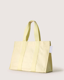The graf lantz light yellow cotton tote bag features two sturdy handles, vertical stitching, and a small white tag on the side. Its neutral grey-beige background makes it perfect for beach days.