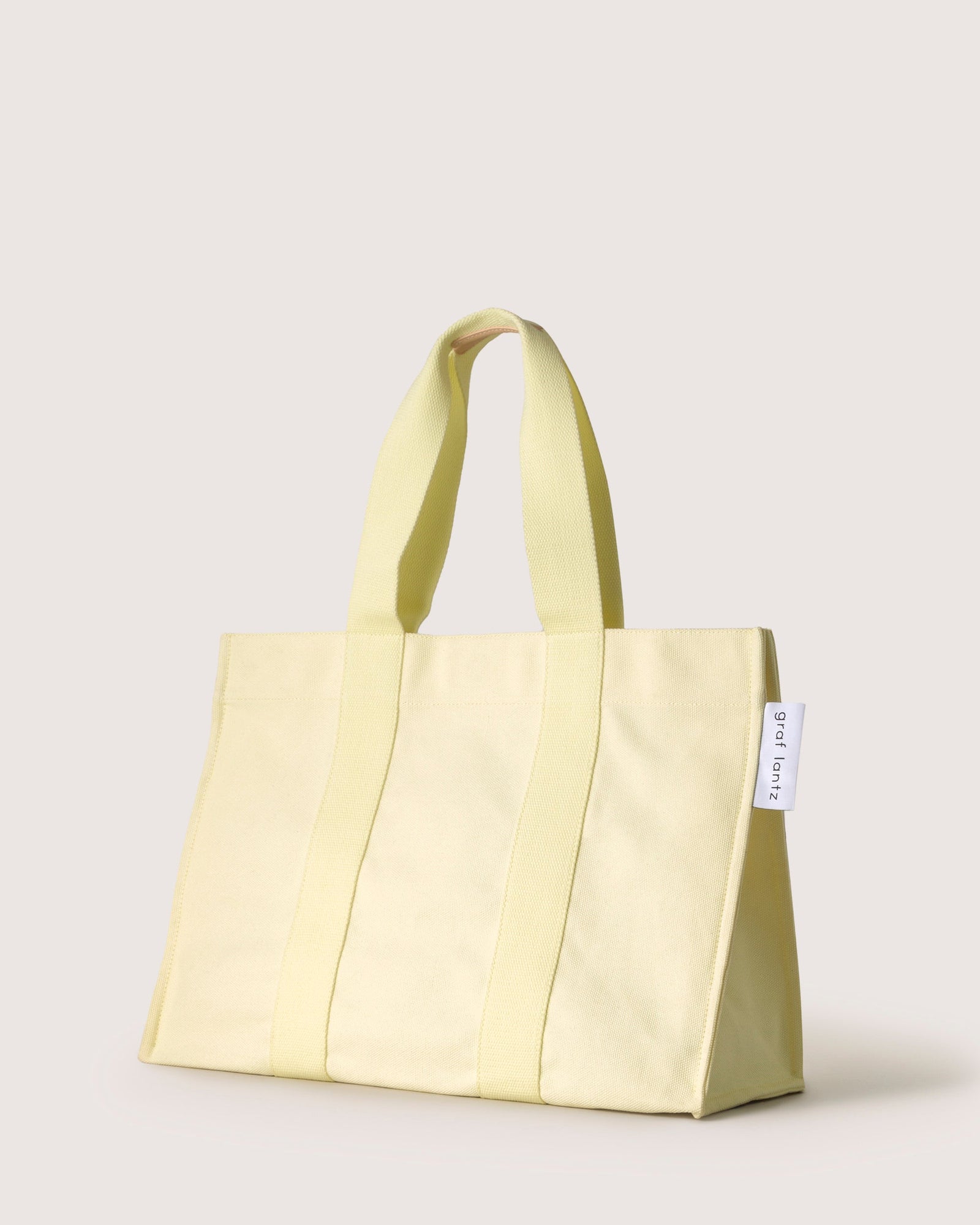 The graf lantz light yellow cotton tote bag features two sturdy handles, vertical stitching, and a small white tag on the side. Its neutral grey-beige background makes it perfect for beach days.