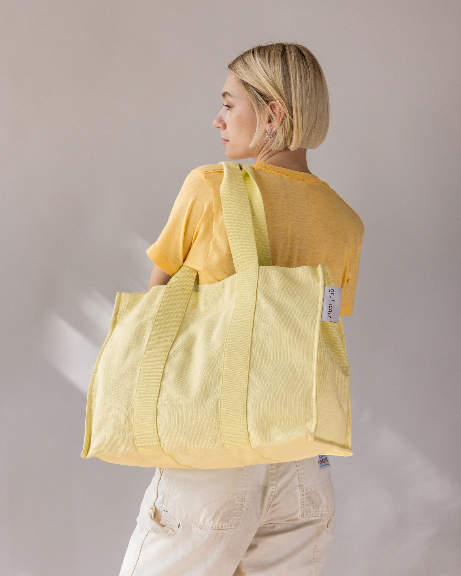 A person with short blonde hair, dressed in a yellow shirt and beige pants, effortlessly carries a large cotton tote from graf lantz with a magnetic closure over their shoulder. The neutral gray backdrop perfectly highlights this beach-ready ensemble.