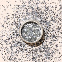 A round container of Manucurist Glitters Duo Silver  Mirror is displayed on a light surface, with its silver and mirror glitters shimmering in a circular pattern around it.